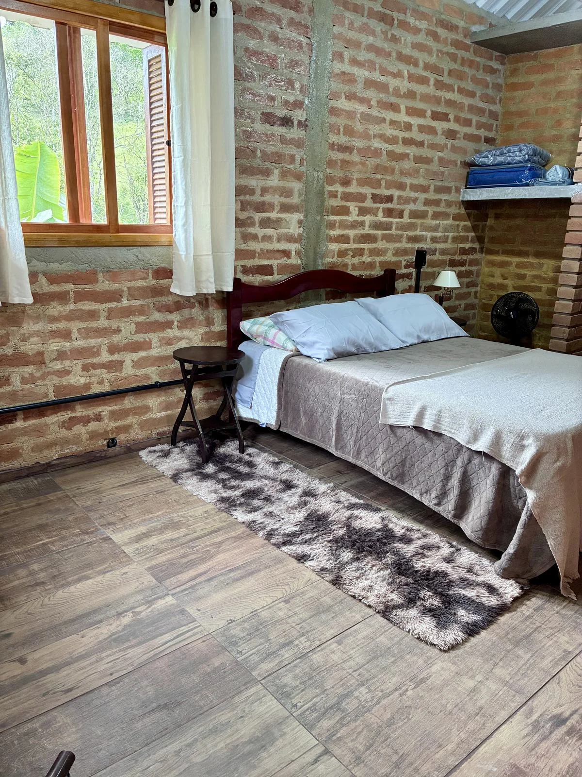 Suíte com cama de casal, janela de madeira e paredes de tijolos